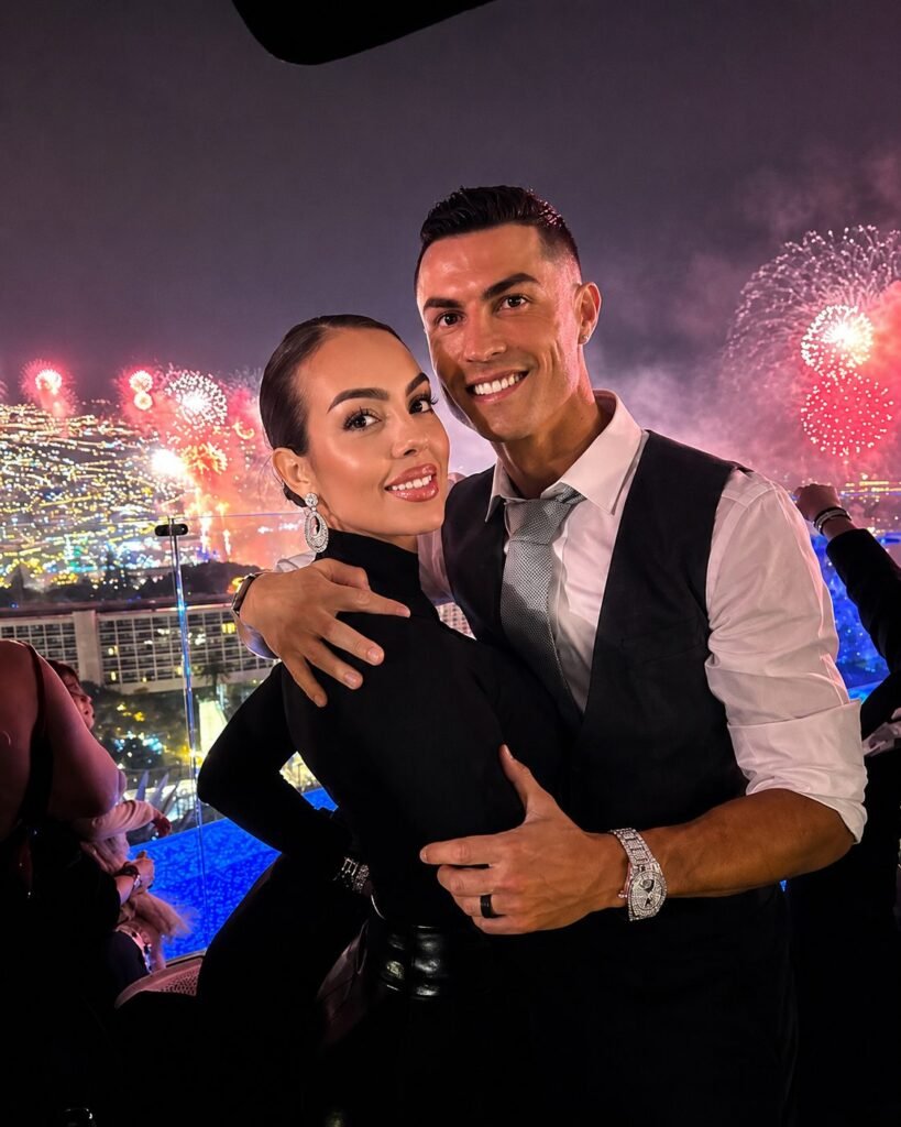 Cristiano Ronaldo and Georgina Rodriguez stand close together on a terrace at night, fireworks bursting over a lit bay behind them.