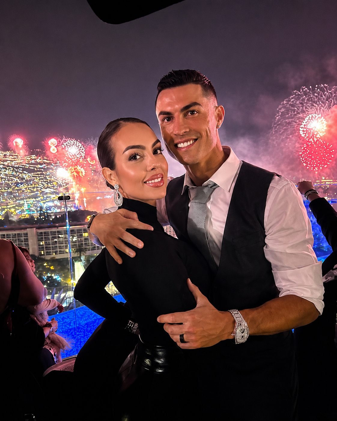 Cristiano Ronaldo and Georgina Rodriguez stand close together on a terrace at night, fireworks bursting over a lit bay behind them.