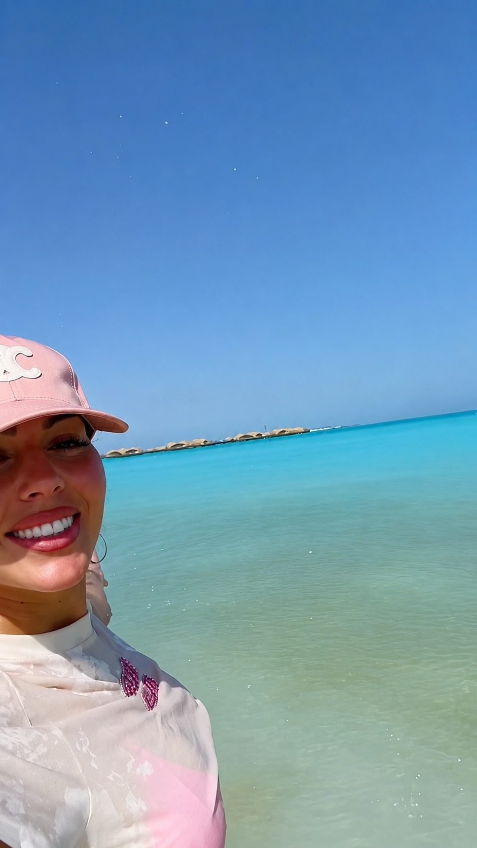 Georgina Rodriguez standing in shallow turquoise water under a clear blue sky, wearing a pink cap.
