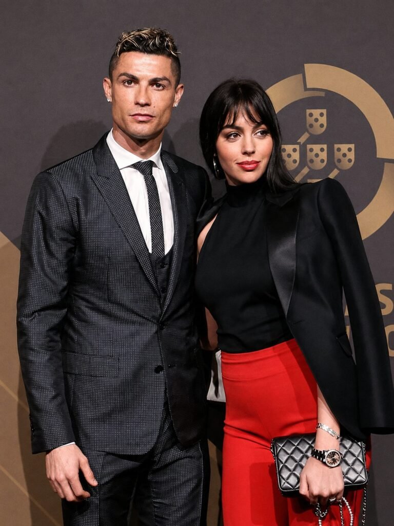 Cristiano Ronaldo in a dark three-piece suit beside Georgina Rodriguez in a black high-neck top and red trousers, against a gold crest backdrop.