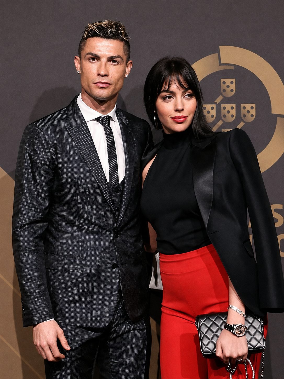 Cristiano Ronaldo in a dark three-piece suit beside Georgina Rodriguez in a black high-neck top and red trousers, against a gold crest backdrop.