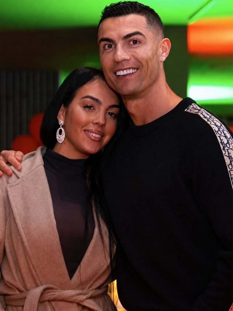 A close portrait of Georgina Rodriguez resting her head against Cristiano Ronaldo cheek, green and amber neon light behind them.