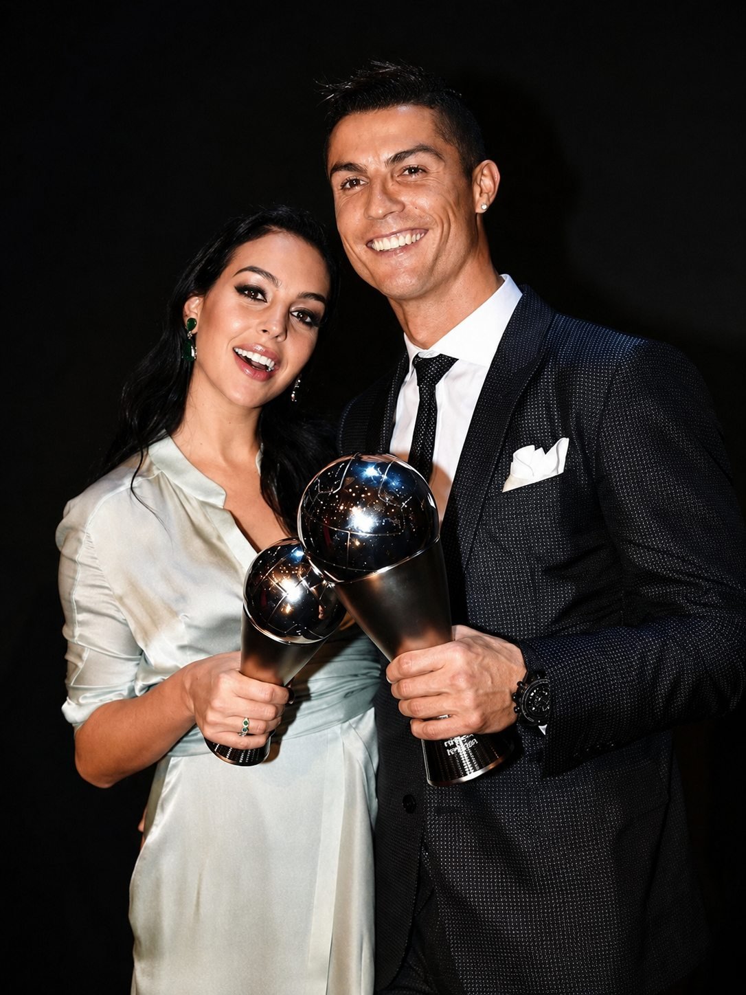 Cristiano Ronaldo and Georgina Rodriguez each holding a football trophy against a black backdrop.