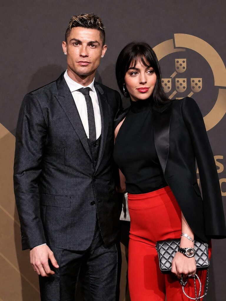 A full-length photograph of Cristiano Ronaldo and Georgina Rodriguez on a red carpet in front of a large gold football crest.