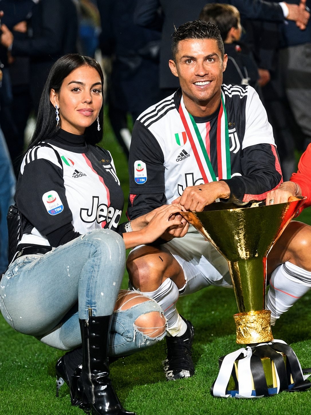 Cristiano Ronaldo and Georgina Rodriguez crouching on a football pitch beside a large gold trophy, both wearing black and white striped Juventus shirts.