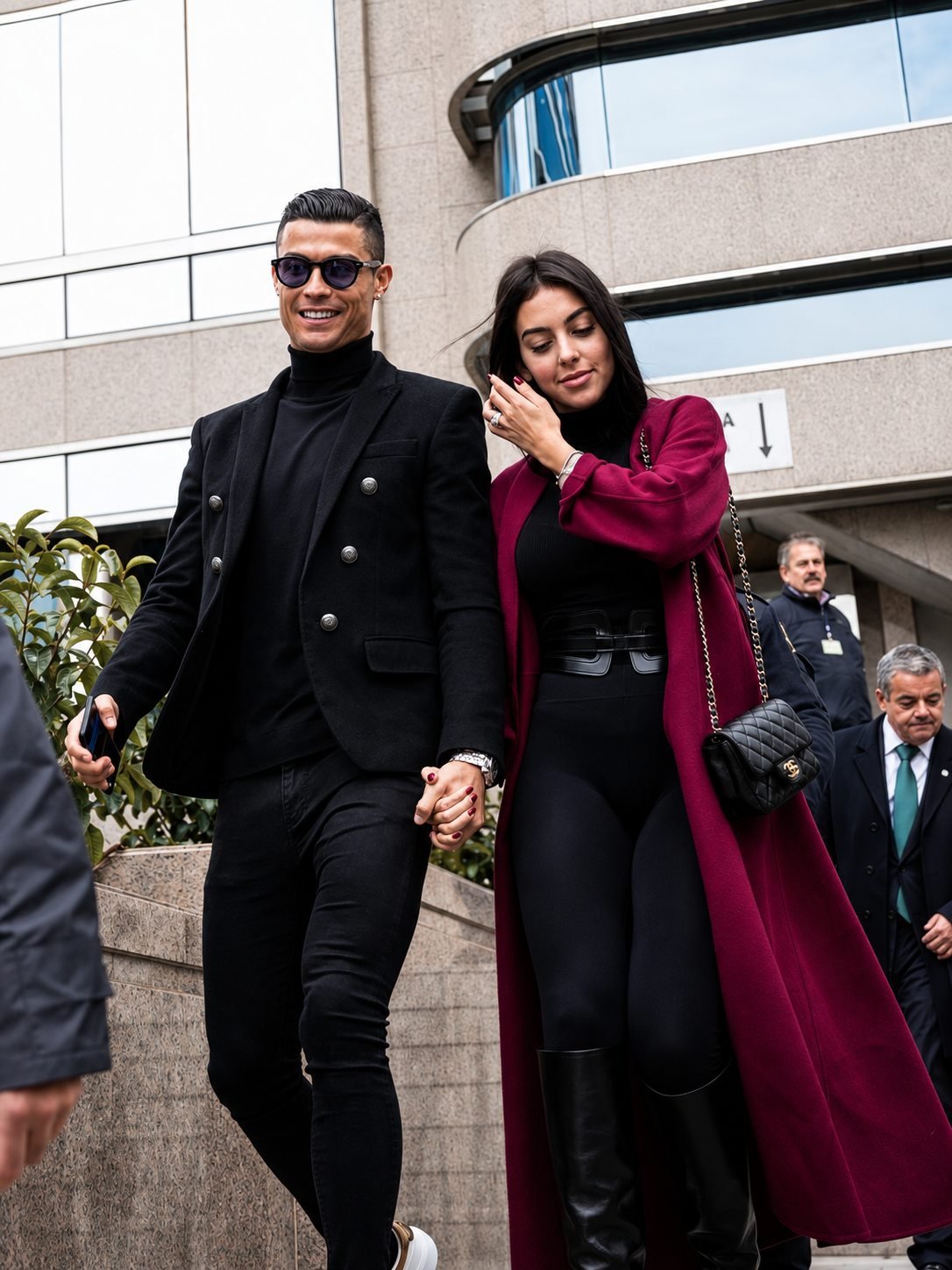 Cristiano Ronaldo and Georgina Rodriguez walking hand in hand down steps outside a stone building, she in a floor-length burgundy coat.